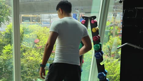 a man in a gym selects dumbbells near a large window with a city view. bright natural lighting enhances the scene