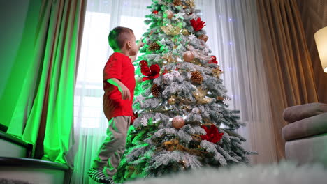 Child gazing at a Christmas tree. A child in a red shirt gazes at a bright Christmas tree in a cozy living room