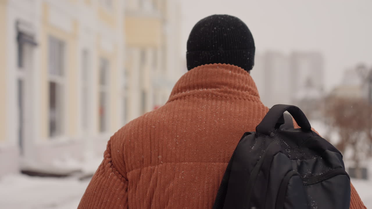 Black man with backpack walks along snowlined avenue past pastel facades and shopfronts, flakes settling on beanie and coat frames hint at designer hurrying to studio, delivery rider weaving, tourist