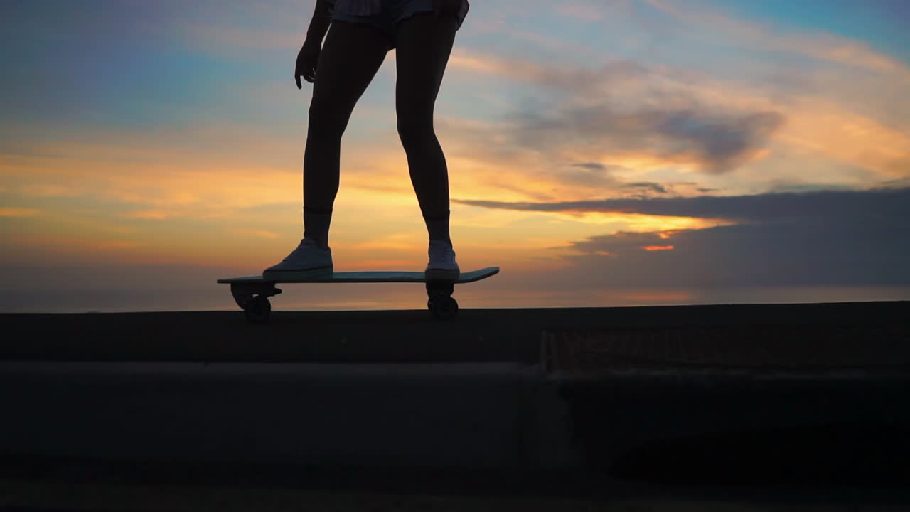 A slow-motion scene captures the essence of two friends skateboarding on a road at sunset, framed by mountains and a captivating sky. Shorts are their chosen attire