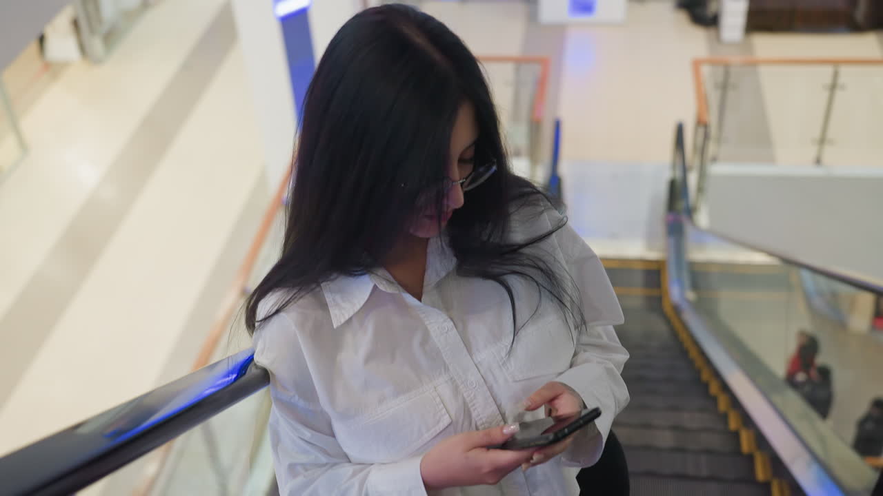 Calm young woman in white shirt and glasses gently leans on escalator railing while descending with phone in hand, appearing relaxed and focused inside bright modern shopping center interior