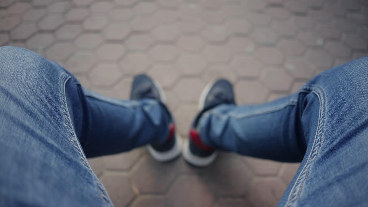 Vista dall'alto di un uomo seduto all'aperto che mostra le gambe in jeans e scarpe da ginnastica sul marciapiede
