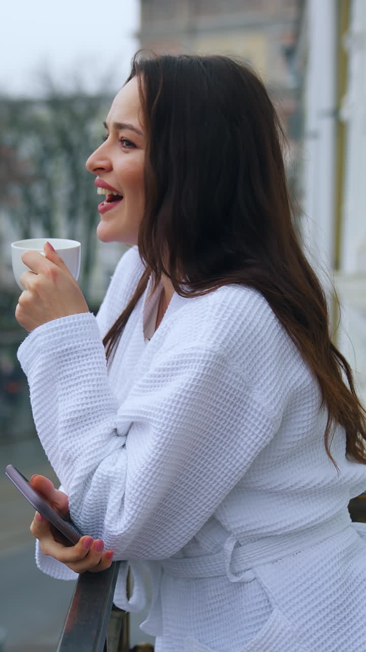 Beautiful young girl in a snow-white terry bathrobe on a balcony with the cup of tea in the morning. Happy holidays away from home. Woman in hotel. Vertical video