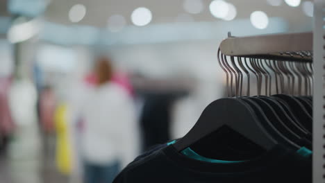 close-up of black hangers holding clothing items in retail store, with blurred background featuring a shopper in white browsing colorful displays