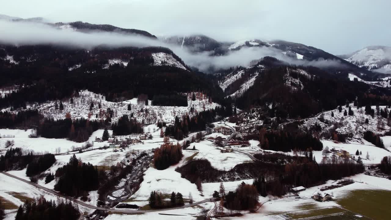 Aerial drone view of snow-covered valley with scattered houses, dense forests, and hills under low clouds covering mountain peaks in Trentino-Alto Adige