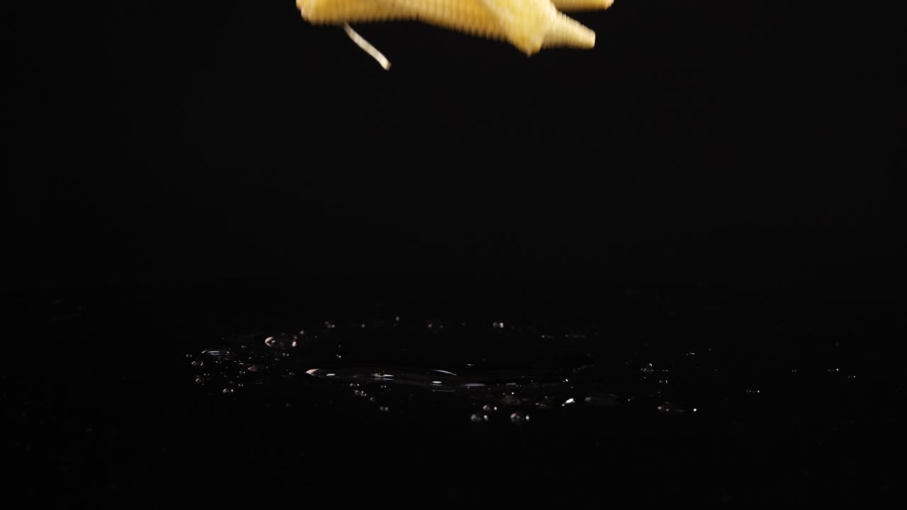 Baby corn pieces drop and settle on a reflective black surface, captured in slow motion with dramatic lighting