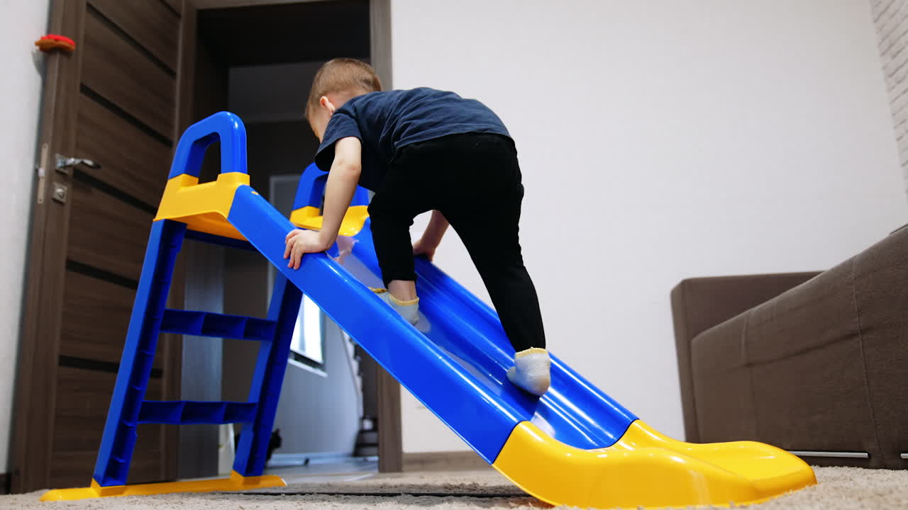 Caucasian toddler climbing the plastic slide. Kid slips down and laughs cheerfully. Low angle view.
