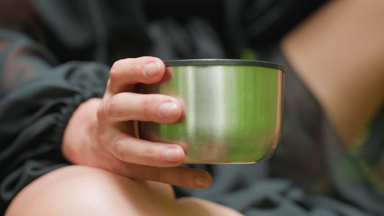 Close up of person holding stainless cup outdoors, dressed in black outfit with soft blurred background and subtle light reflections, creating minimalist mood, tactile realism