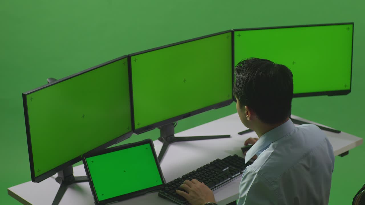 vista lateral de un hombre asiático trabajando con una tableta y varios monitores de computadora con una maqueta de pantalla verde en un estudio de pantalla verde