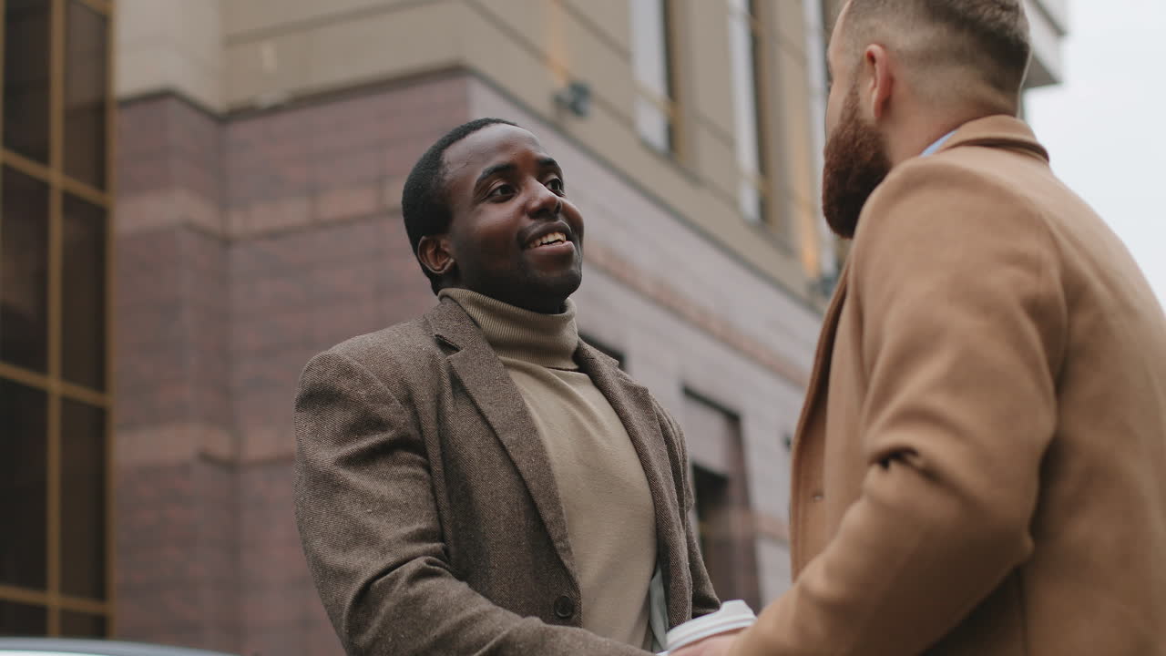 hombre de negocios afroamericano hablando por teléfono y mirando el cuaderno abierto en sus manos en la calle en otoño, luego un hombre caucásico se acerca a él y le da la mano