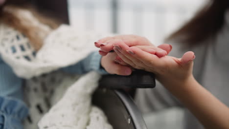 Mother-strokes-hand-of-little-daughter-sitting-in-wheelchair