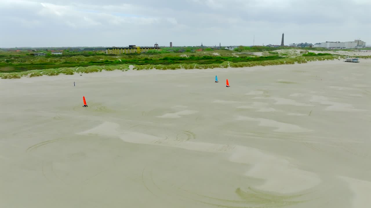 Colorful land yachts glide across the vast flat beach of Borkum Island. Grassy dunes and distant resort buildings in the background