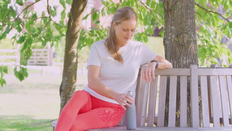 woman wearing fitness clothing sitting on seat under tree checking activity monitor on smartwatch