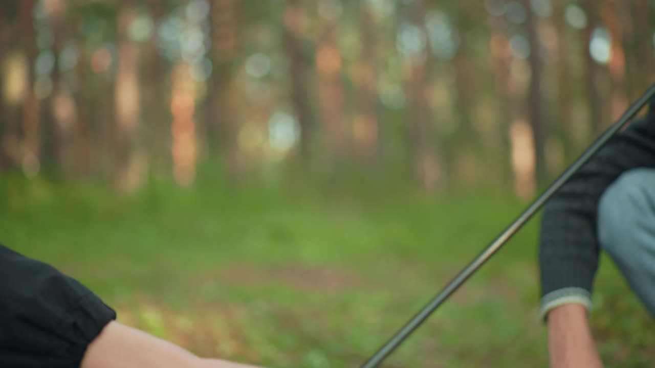 Partial view of man squatting while assembling tent pole during forest camping as another person reaches out to hand over supporting pole, creating a teamwork moment in outdoor nature setting