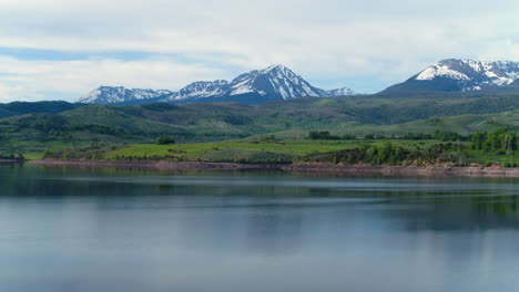 Aerial-Drone-of-Lake-in-Colorado-Rocky-Mountains-with-Forest-Trees-and-Snow-Capped-Peaks-in-the-Distance