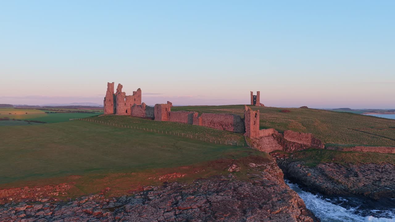Drone flyver over Dunstanburgh slotsruiner ved solopgang
