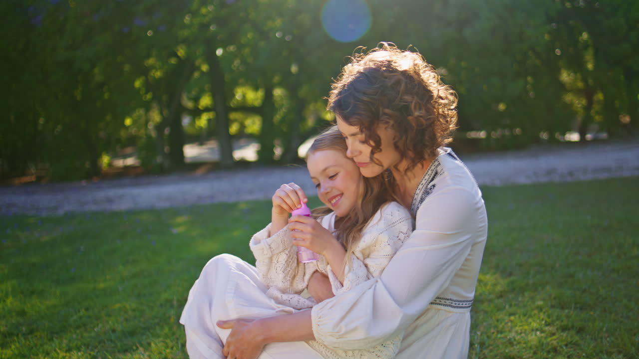 Happy family blowing soap bubbles sitting on green grass in sunny park closeup