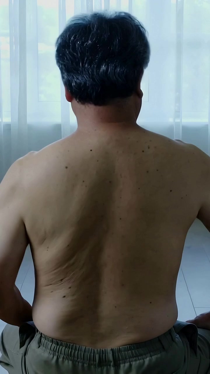 Video verticale: Uomo a torso nudo che cambia posizione, aggiusta la postura e ruota la testa in una stanza illuminata dal sole con una sedia.