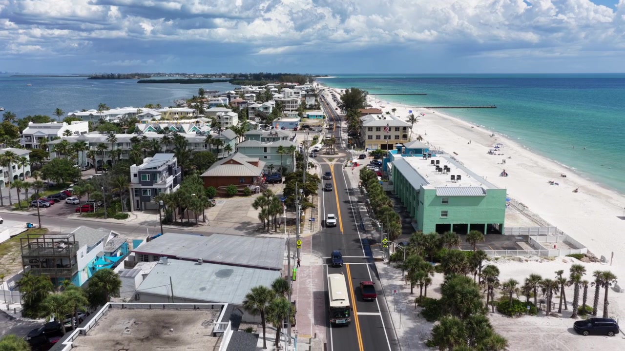 La strada costiera di Bradenton, Florida, si snoda tra colorati edifici pastello e palme ondeggianti, con auto che viaggiano verso un'ampia costa sabbiosa lambita dalle acque turchesi del golfo. Ripresa aerea con drone all'indietro.