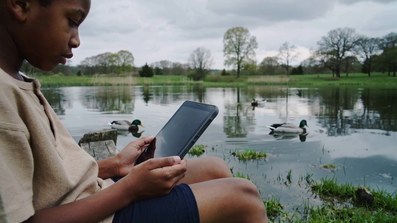 Adjusting boy tan tee navy shorts tapping tablet park pond mallards approach prompt lean