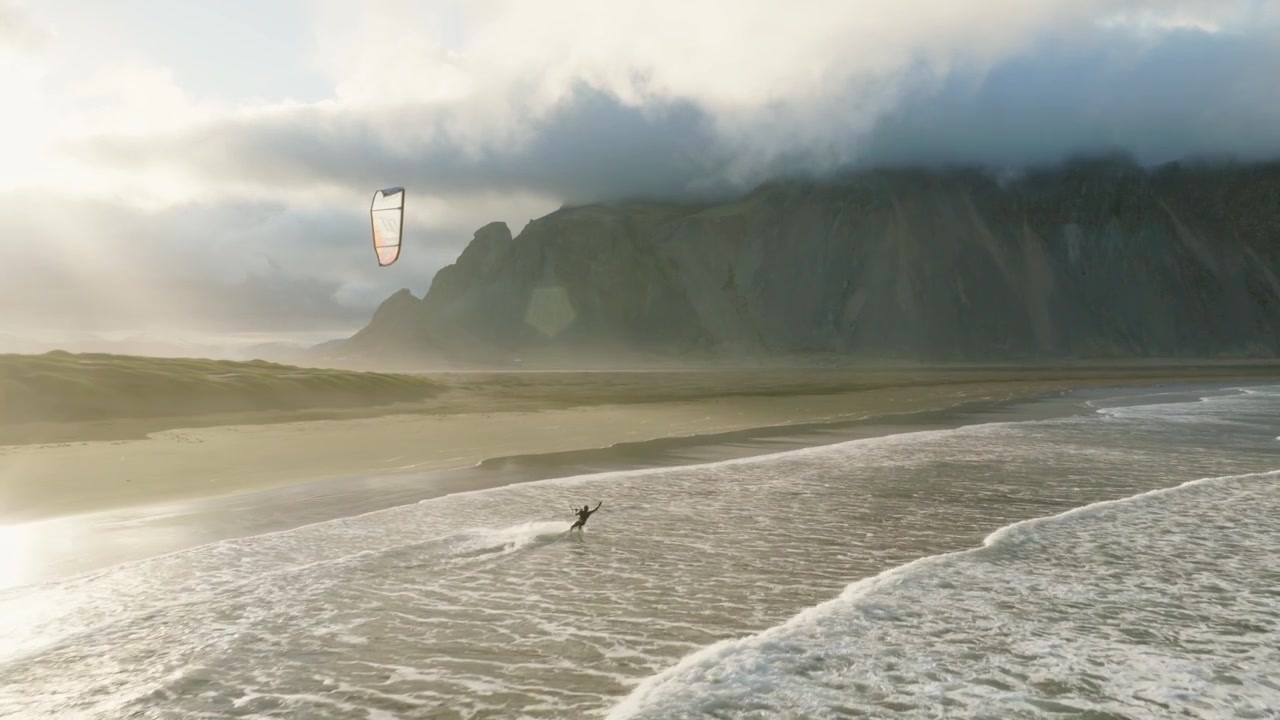 Kite surfer follows Atlantic shoreline beneath distinctive Vestrahorn mountain ridge, with dark coastal sand, breaking waves and low cloud covering upper slopes at Stokksnes, Southeast Iceland, aerial