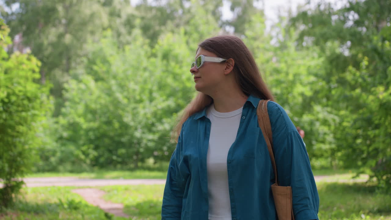 Woman in blue shirt and white sunglasses stands in green park admiring nature while person passes by in soft blur background, sunlight filtering through trees creating calm serene summer atmosphere