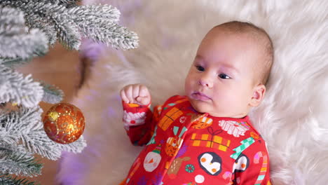 calm peaceful baby boy in red shirt lies on soft plaid under new year tree. sweet child looks up at shiny decoration ball hanging over him. view from above.
