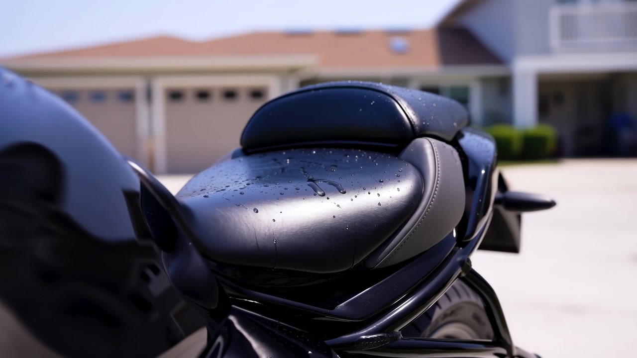 Närbild av svart läder passagerarsäte på motorcykel med litet vadderat ryggstöd, pärlande droppar, tunna avrinningslinjer, glansiga reflektioner och en förortsuppfart bredvid ett hus i ljust