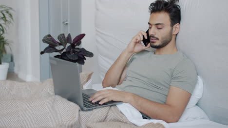 man working from bed with laptop and phone