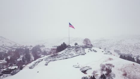Aerial-shot-of-the-American-flag-proudly-waving-on-top-of-a-snowy-peak