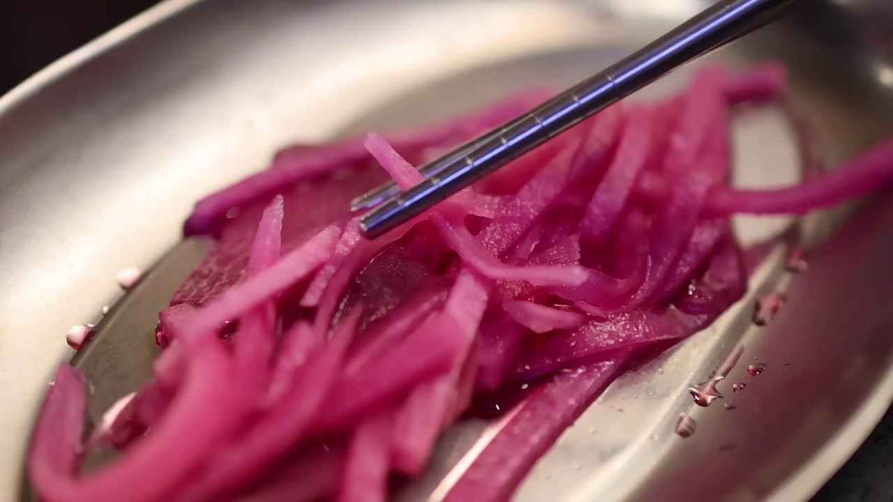 Close-up of chopsticks picking up bright pink pickled onions from a stainless steel dish.