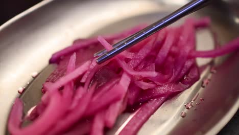 close-up of chopsticks picking up bright pink pickled onions from a stainless steel dish.
