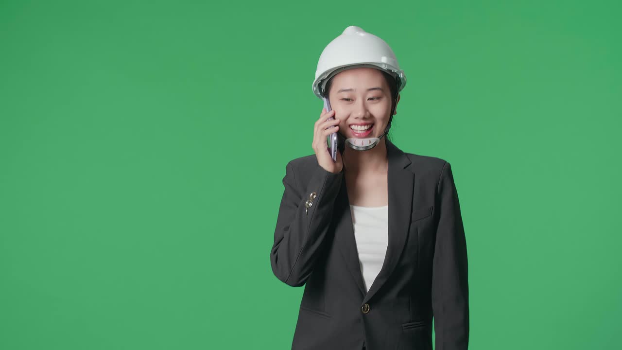 Smiling Asian Female Engineer With Safety Helmet Talking On Smartphone And Walking In The Green Screen Studio