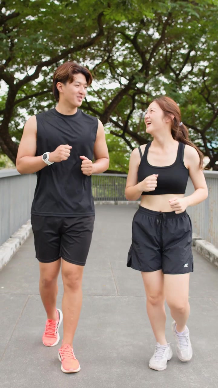 Coppia asiatica sorridente fa jogging insieme su un ponte di un parco alberato, con movimento di telecamera a mano con tracciamento in avanti
