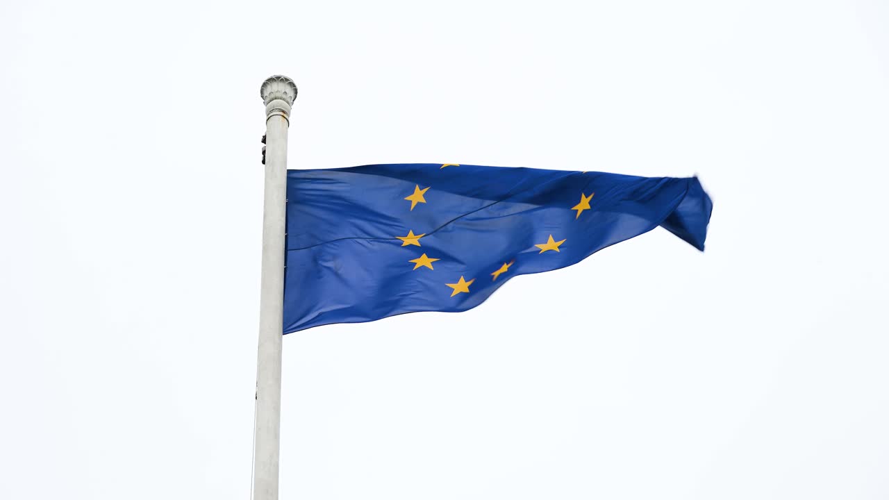 Slow-motion shot of the European Union (EU) flag fluttering on a large post, swaying in the wind on a cloudy, breezy day, emphasizing the flag's fluid motion against the elements.
