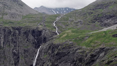 Toma-Aérea-Con-Plataforma-Rodante,-Avanzando-Lentamente-Sobre-Un-Río-Largo-Y-Delgado,-Serpenteando-Por-Una-Ladera-Empinada,-Verde-Y-Cubierta-De-Musgo-Junto-A-Los-Trollstigen,-Noruega