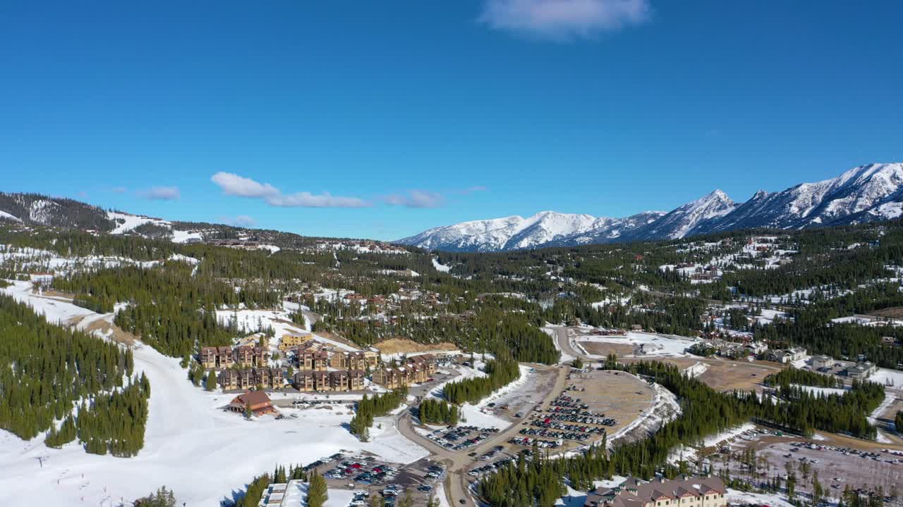Drone soars above a busy winter parking lot near the heart of Big Sky, with snow-draped condos, icy roads, and distant peaks just steps from the base of the mountain resort