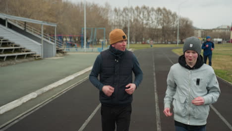 close up of a coach jogging alongside with a young boy on a track in a stadium with someone also coming from behind jogging as the coach talks to the boy