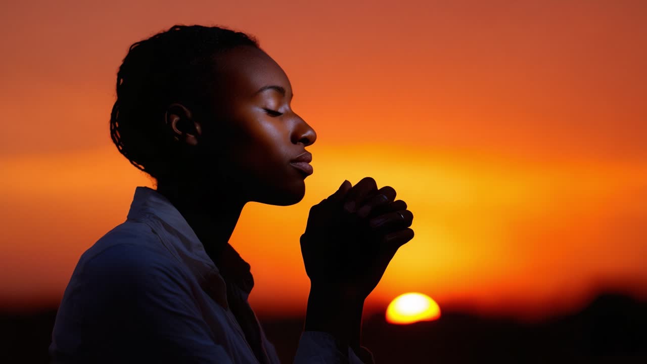 Une femme aux yeux fermés et aux mains jointes prie pendant un coucher de soleil à couper le souffle, vivant un moment de profonde réflexion et de connexion avec son environnement et ses pensées.