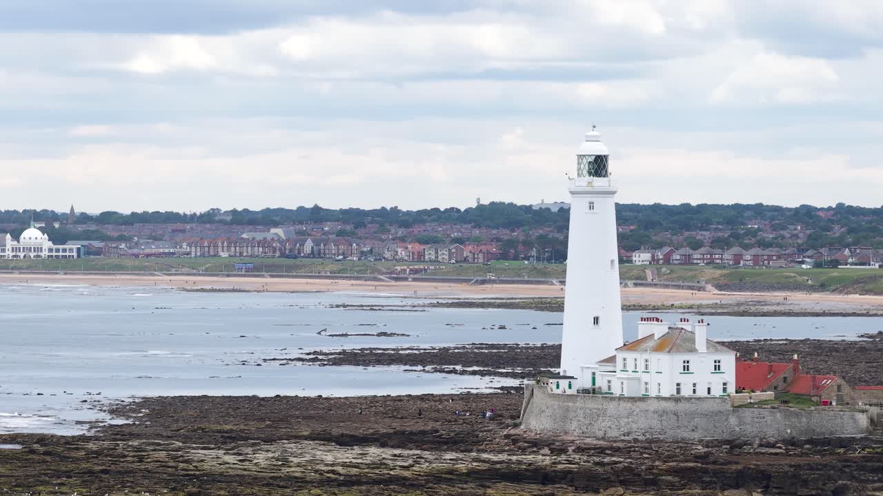 A steady side-to-side camera pan reveals St Mary’s Lighthouse on a rocky tidal shore under overcast daylight, with distant coastal town in background