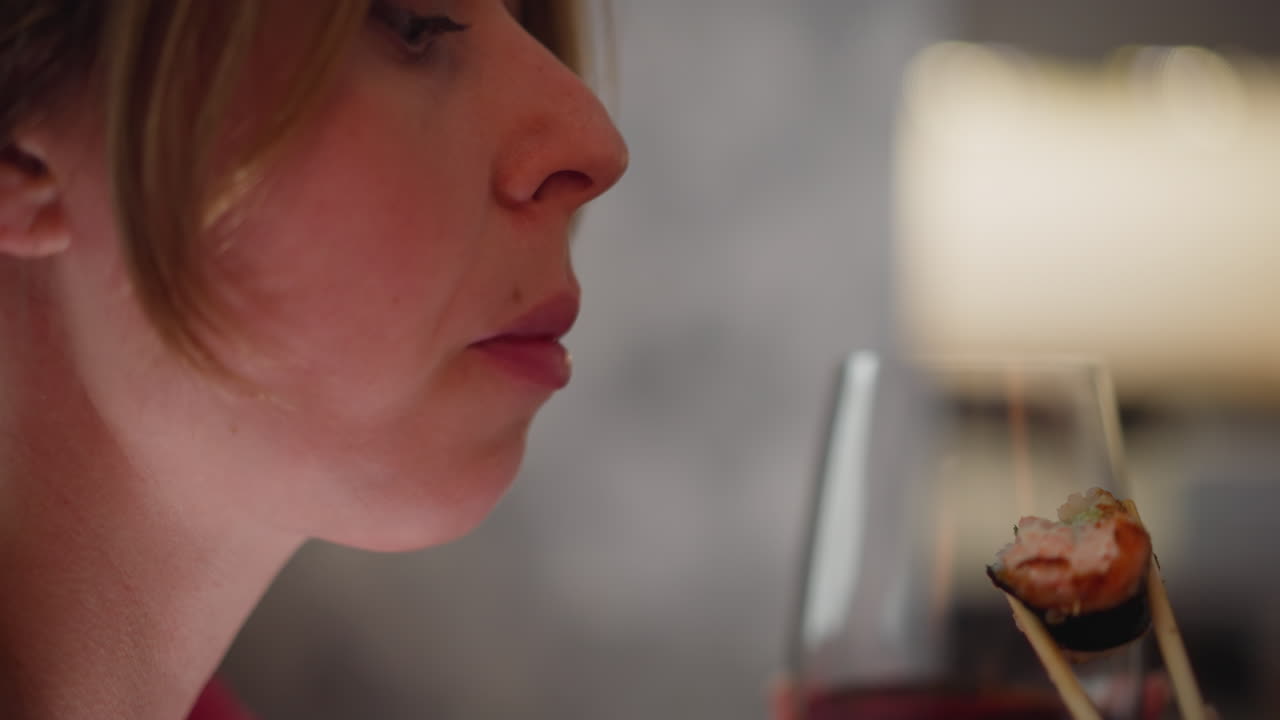 Close up of girl licking sushi sauce with tongue as she eats sushi using chopstick while holding glass of red wine showing expression of enjoyment and focus in cozy indoor setting