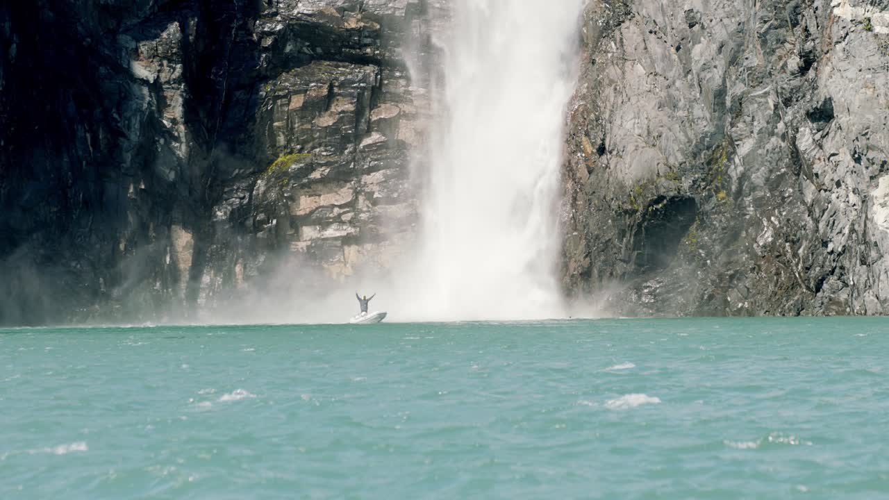 Happy Male in a Motorboat Under Glacial Waterfall in Wilderness of Alaska, Slow Motion. Exploring Alaskan Nature on Sunny Day