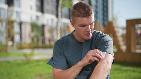 young man seated outdoors applying nicotine patch to upper arm with modern residential complex blurred background casual clothing focused expression sunlight greenery wooden park bench