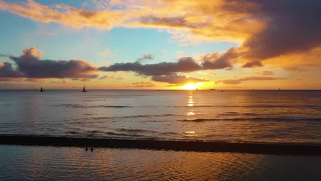 golden hour sunrise over ocean in waikiki beach, hawaii as waves crash into breakwall with sailboats and people