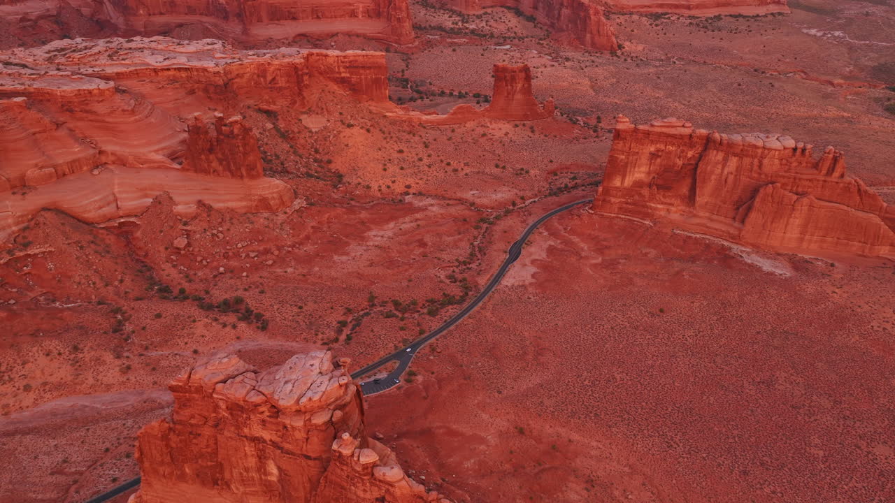 Lonely white car moves by the modern highway crossing the red arid desert. Aerial view on the spectacular canyons of the Arches National Park, Utah, USA at sunset