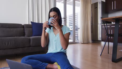 Mixed-race-gender-fluid-person-sitting-on-floor-drinking-coffee-and-using-laptop-at-home