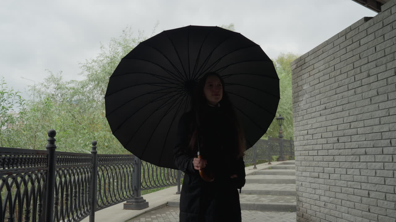 Donna che cammina con un ombrello nero lungo la passeggiata del fiume; il cielo nuvoloso e il marciapiede bagnato creano un'atmosfera malinconica; ringhiera in ghisa, lampioni vintage, muro di mattoni e alberi distanti incorniciano la scena solitaria