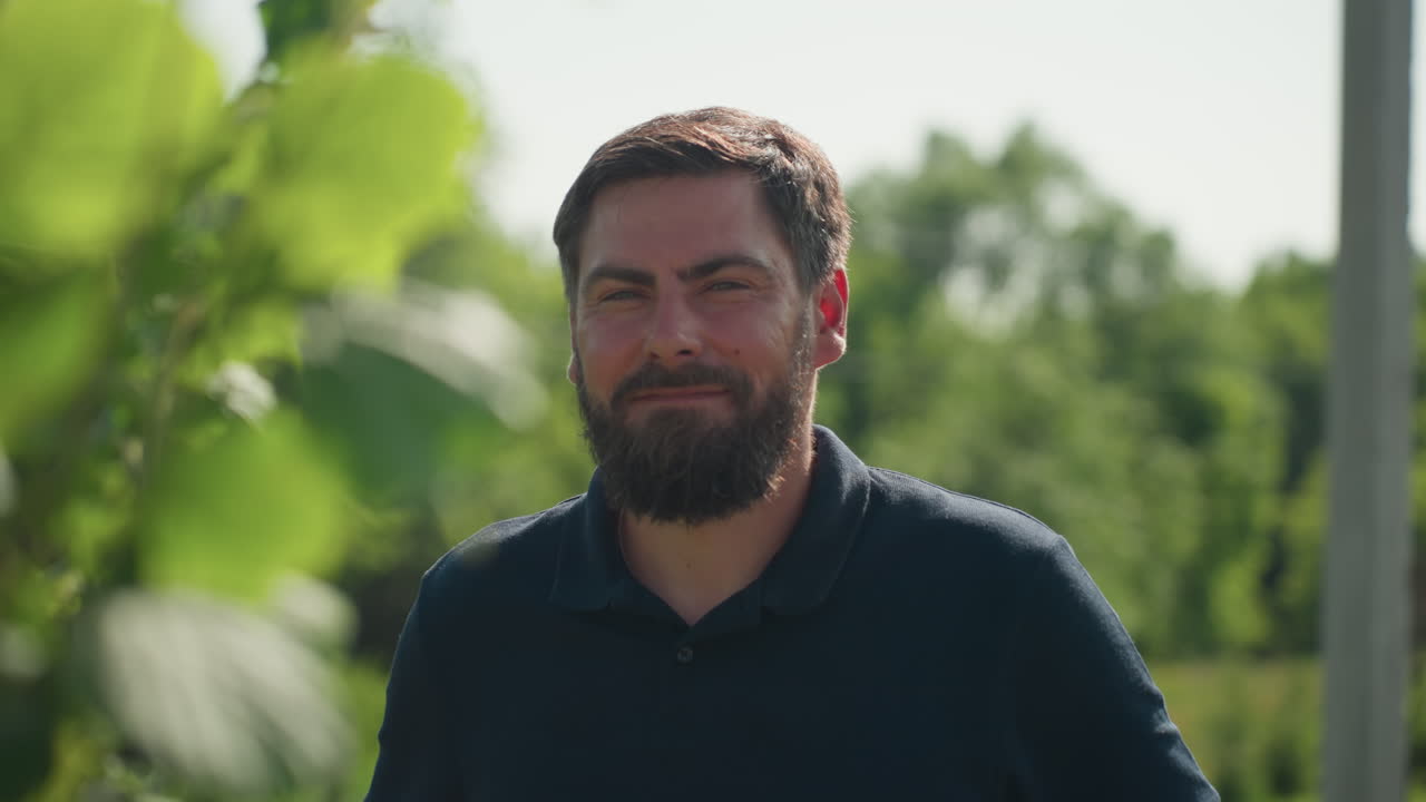 farmer turns with calm expression among green leaves, side profile framed by blurred foliage, sunlight highlighting beard and shirt, peaceful rural garden scene with gentle breeze