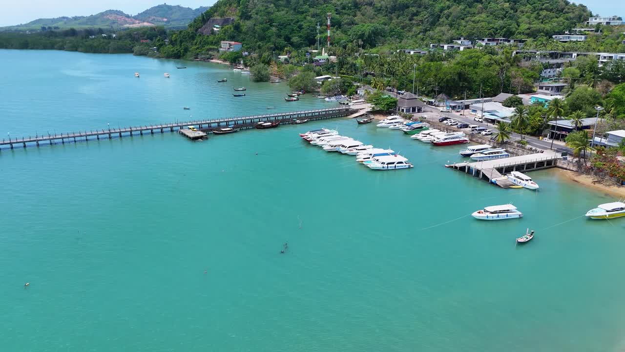 Drone footage captures a serene marina in Phuket, Thailand, showcasing boats, clear waters, and lush greenery under bright daylight