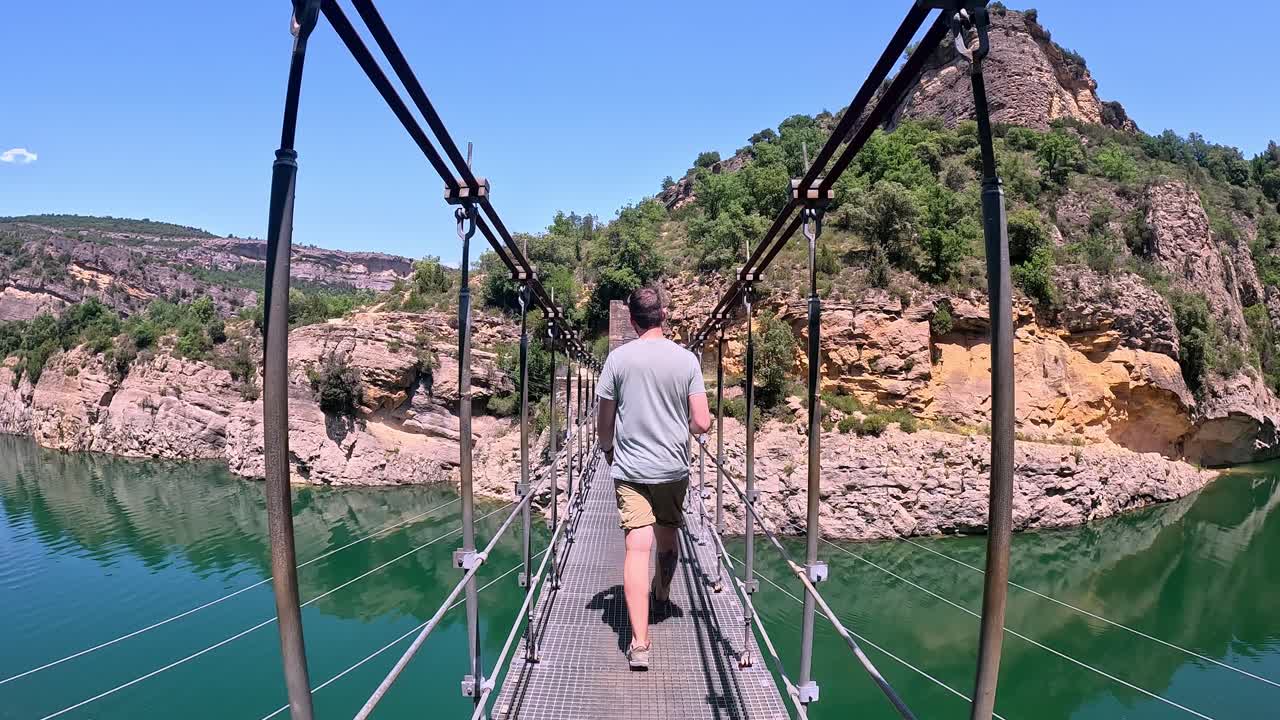 A back view of a young man wearing casual attire, confidently walking across a scenic hanging bridge over the turquoise waters of Congost de Mont Rebei.